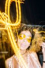Самоуверенная молодая афроамериканка с вьющимися волосами в модных солнцезащитных очках и топ расслабляющая на улице вечером возле замораживающих огней — стоковое фото