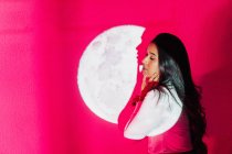 Vista laterale della giovane donna ispanica in piedi con gli occhi chiusi toccando guance sotto brillante illuminazione al neon rosa e proiezione lunare — Foto stock