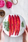 Top view of crop unrecognizable person showing gravlax with mixed peppercorns and fresh dill sprigs on plate on light background — Stock Photo
