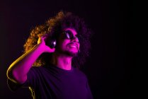 Delizioso maschio latino con capelli ricci in occhiali da sole e cuffie che ascolta musica in studio con luci al neon su sfondo nero — Foto stock