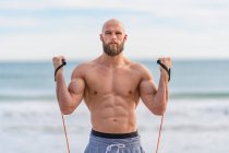 Shirtless brutal man looking at camera doing side lateral flies with resistant band training on seashore and looking forward — Stock Photo