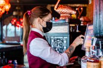 Вид збоку жінки-банкіра в захисній масці з використанням касової коробки та сенсорного екрану дисплея під час роботи в барі під час коронавірусу — стокове фото