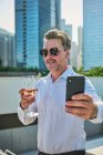 Designer après le travail ayant du temps de détente dans une terrasse à côté des immeubles de bureaux, il prend une tasse de vin blanc tout en prenant un selfie — Photo de stock