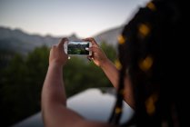 Cultivo irreconocible viajera afroamericana tomando fotos en el teléfono inteligente del estanque en las tierras altas - foto de stock