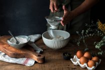 Неузнаваемый человек просеивает муку в миску во время приготовления выпечки на деревенской кухне дома — стоковое фото