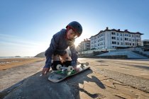 Cheerful teenage boy in helmet sitting with skateboard on promenade near sea in summer — Stock Photo