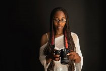 Serious African American female photographer with braids looking through photos taken on professional camera while standing on black background — Stock Photo