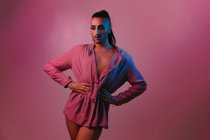 Porträt einer glamourösen Transgender-bärtigen Frau in raffiniertem Make-up, die mit den Händen auf der Taille vor rosa Hintergrund im Studio posiert und wegschaut — Stockfoto