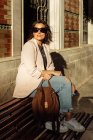 Corpo inteiro de mulher na moda em roupa casual elegante e óculos de sol com bolsa sentada no banco e desfrutando da luz solar enquanto descansa na rua urbana — Fotografia de Stock