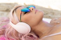 Жінка з рожевим волоссям і в сонцезахисних окулярах лежить на рушнику на піщаному березі і розслабляється під час літніх канікул, слухаючи музику в навушниках — стокове фото