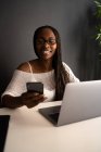 Allegra freelance afroamericana seduta a tavola sul posto di lavoro moderno e che naviga sul cellulare mentre lavora su un progetto remoto da casa — Foto stock