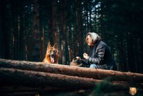 Vista lateral do homem sentado em um tronco de árvore perto de árvores coníferas com cão doméstico no dia ensolarado — Fotografia de Stock