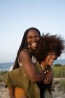 Cheerful young African American female with Afro pigtails giving piggyback ride to laughing girlfriend with curly hair while having fun during summer holidays together on seashore — Stock Photo