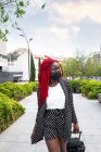 Jeune femme afro-américaine en masque de protection avec dreadlocks lumineux flânant dans le parc avec valise et détournant les yeux — Photo de stock