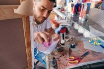 High angle of male artist pouring liquid pigment into spray gun while sitting near table behind easel with canvas in workshop — Stock Photo