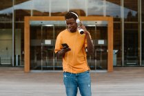 Fröhlicher afroamerikanischer Mann surft auf Smartphone und hört Musik über Kopfhörer, während er gegen ein modernes Gebäude läuft — Stockfoto