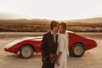 Glückliche Männer und Frauen in eleganten Kleidern, die sich bei der Hochzeitsfeier im Bardenas Reales Naturpark in Navarra, Spanien, in der Nähe eines Sportwagens vor dem Sonnenuntergang umarmen — Stockfoto