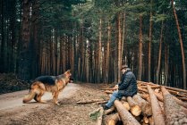 Vue latérale de l'homme assis dans un tronc d'arbre près des conifères avec chien domestique dans la journée ensoleillée — Photo de stock