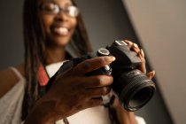 Fotógrafa afroamericana con trenzas mirando a través de fotos tomadas en cámara profesional mientras está de pie sobre fondo negro - foto de stock