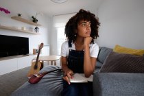 Nachdenkliche afroamerikanische Musikerin sitzt mit Mandoline und Notizbuch im Bett, während sie Lieder komponiert und zu Hause wegschaut — Stockfoto