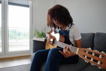 Talentuosa musicista afroamericana che suona la chitarra acustica seduta sul divano di casa — Foto stock