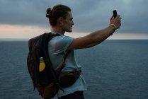 Viajero masculino de vista lateral tomando una foto del cielo en el teléfono inteligente en verano - foto de stock