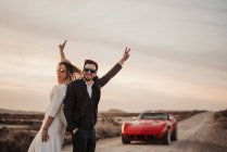 Optimistic bride and groom gesturing V sign and smiling while standing on road near red car in evening in Bardenas reales Natural Park in Navarra, Spain — Stock Photo