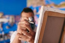 Recorte artista masculino irreconocible en respirador utilizando pistola de pulverización para pintar cuadro sobre lienzo durante el trabajo en taller creativo - foto de stock