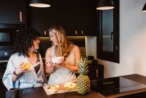 Высокий угол веселой лесбийской пары счастливо смеются, когда режут ананас и едят хлопья с ягодами и йогуртом — стоковое фото