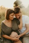 Задоволений чоловік обіймає вагітної жінки закритими очима, сидячи на лузі в сільській місцевості — стокове фото