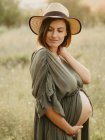 Спокойная беременная женщина в платье и соломенной шляпе трогает животик, стоя на поле в сельской местности на закате летом, глядя в сторону — стоковое фото