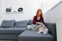 Жінка з іграшкою для домашніх тварин розважається з чарівною пухнастою кішкою, сидячи на дивані вдома — стокове фото