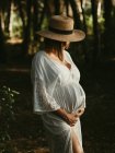 Heitere schwangere Frau in Kleid und Strohhut, die den Bauch berührt, während sie im dunklen Wald steht und wegschaut — Stockfoto