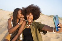 Alegre joven afroamericana amigas sonriendo brillantemente mientras toma selfie en el teléfono inteligente durante el fin de semana de verano en la orilla del mar de arena - foto de stock
