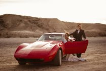 Noiva e noivo descansando perto de veículo vermelho de luxo durante a viagem através do Parque Natural Bardenas Reales de manhã em Navarra, Espanha — Fotografia de Stock