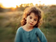 Linda niña étnica de pelo rizado mirando a la cámara contra la borrosa pradera verde al atardecer en verano - foto de stock