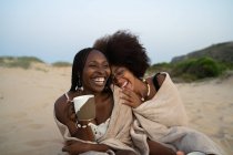 Glückliche junge schwarze Frauen beste Freundinnen mit Bechern mit heißen Getränken sitzen eng in eine warme Decke gehüllt und haben Spaß während des Sommerabends am Sandstrand — Stockfoto
