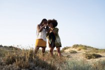 Low angle of cheerful young African American female friends in stylish summer clothes and accessories checking pictures on photo camera while spending summer holidays together in countryside — Stock Photo