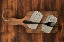 De cima arranhado tábua de corte com garfo e latas seladas com alimentos preservados em mesa rústica madeira — Fotografia de Stock