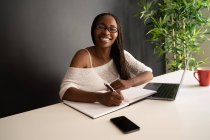 Contenu noir femme pigiste écrivant des notes dans un carnet tout en étant assis à la table avec un ordinateur portable et de travailler sur un projet d'entreprise dans le bureau à domicile moderne — Photo de stock