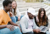 Diversos jovens amigos sorrindo amplamente enquanto assistem vídeo no netbook juntos enquanto estão sentados em escadas de concreto — Fotografia de Stock