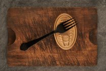 De dessus planche à découper rayée avec fourchette et boîte scellée avec de la nourriture conservée sur table de bois rustique — Photo de stock