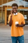 Sonriente hombre afroamericano con taza de café riendo felizmente mientras habla por teléfono móvil y mirando hacia otro lado - foto de stock