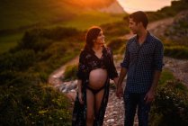 Happy Hispanic pregnant woman and loving man holding hands looking at each other while walking along hilly seashore in summer evening — Stock Photo