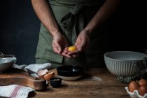 Unrecognizable guy in apron breaking raw egg over bowl while preparing pastry on lumber table near kitchenware — Stock Photo