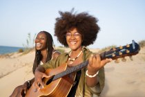 Счастливые молодые афроамериканские подруги играют на гитаре, сидя с закрытыми глазами на песчаном побережье и наслаждаясь летними каникулами — стоковое фото