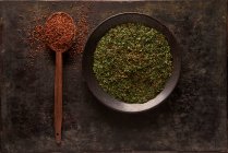 Composition aérienne avec cuillère avec tomates séchées au soleil moulues placées près du bol avec une épice verte à base de plantes sur fond noir — Photo de stock