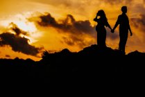 Low angle side view silhouettes of unrecognizable couple expecting baby standing back to back and holding hands while admiring sundown in mountains together — Stock Photo