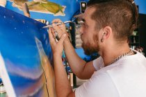 Vista lateral do artista masculino usando pistola de pulverização para pintar quadro em tela durante o trabalho em oficina criativa — Fotografia de Stock