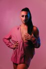 Портрет гламурної трансгендерної бородатої жінки у витонченому макіяжі позує руками на талії на рожевому тлі на студії, дивлячись на камеру — стокове фото